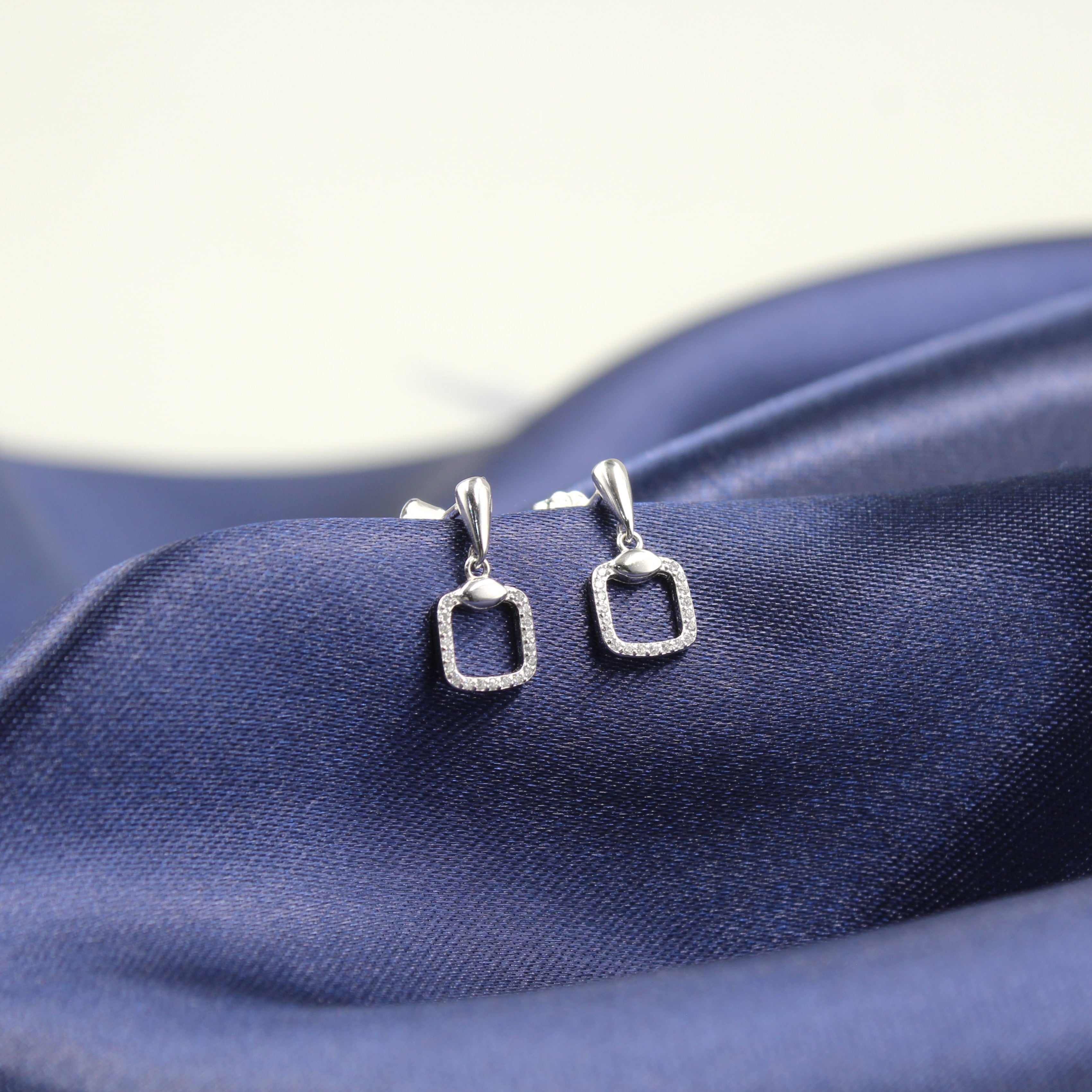 Aretes de plata con forma de rectángulo hueco y borde de diamantes, colocados sobre tela azul.