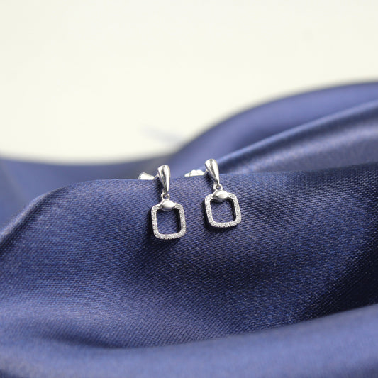 Aretes de plata con forma de rectángulo hueco y borde de diamantes, colocados sobre tela azul.