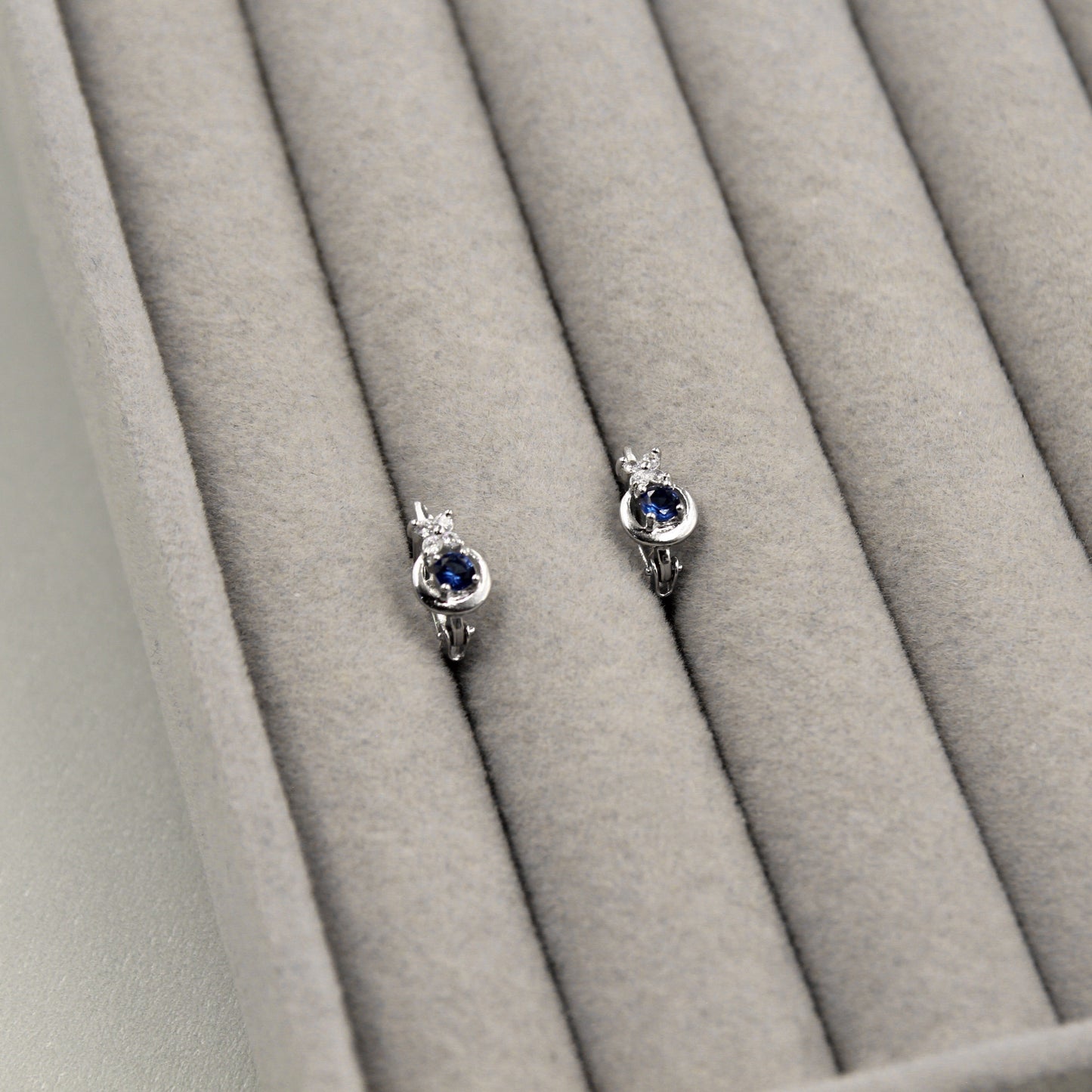 Par de aretes pequeños con zafiros azules redondos, rodeados de un marco de diamantes y un diseño en espiral, presentados en un estuche de joyería gris.