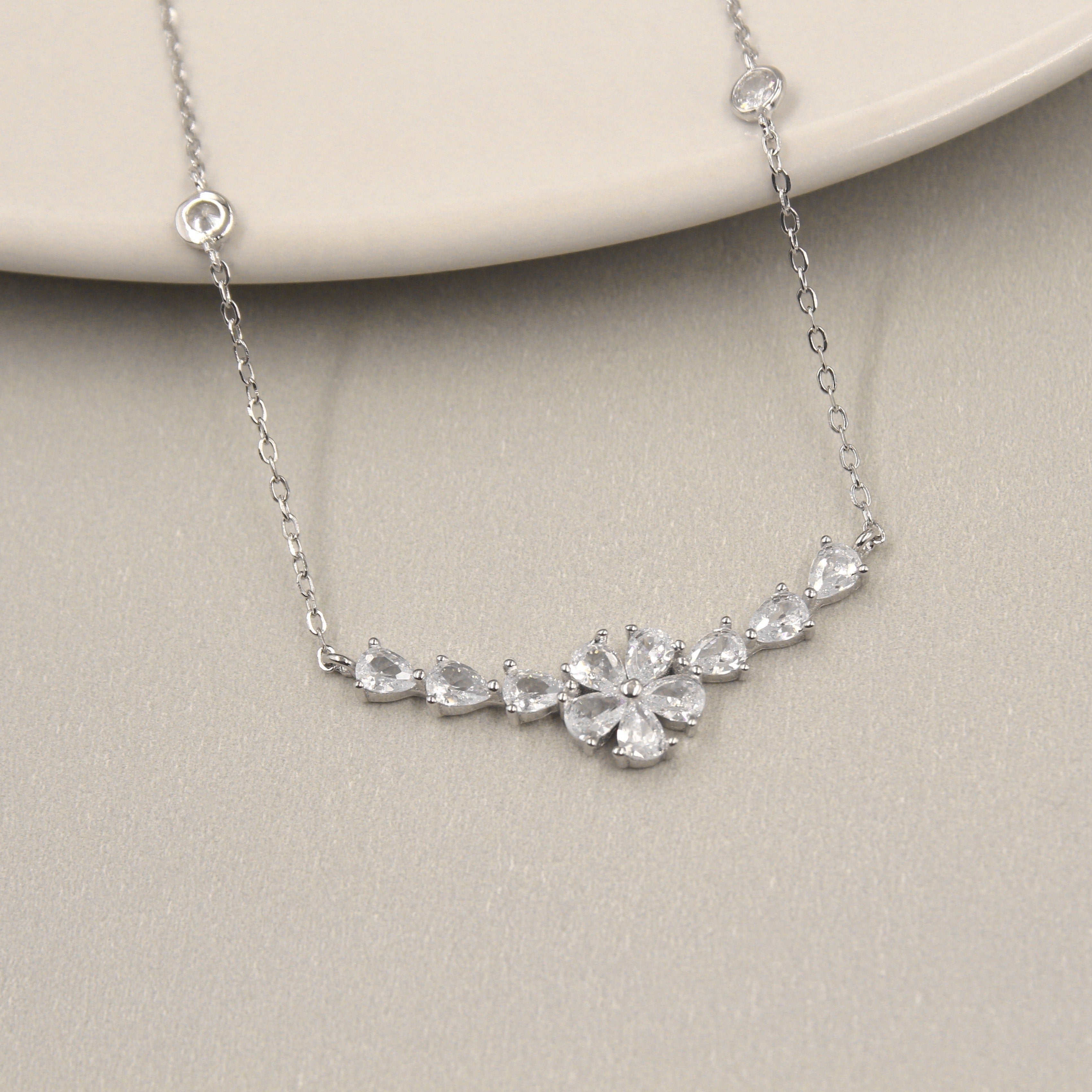 elegante collar de plata con un diseño de colgante que destaca cinco piedras de circonia cúbica dispuestas en un patrón floral. Las piedras varían en tamaño, creando un efecto de flor delicada y radiante. El collar está presentado sobre un plato de cerámica de color claro, resaltando su brillo y diseño sofisticado.