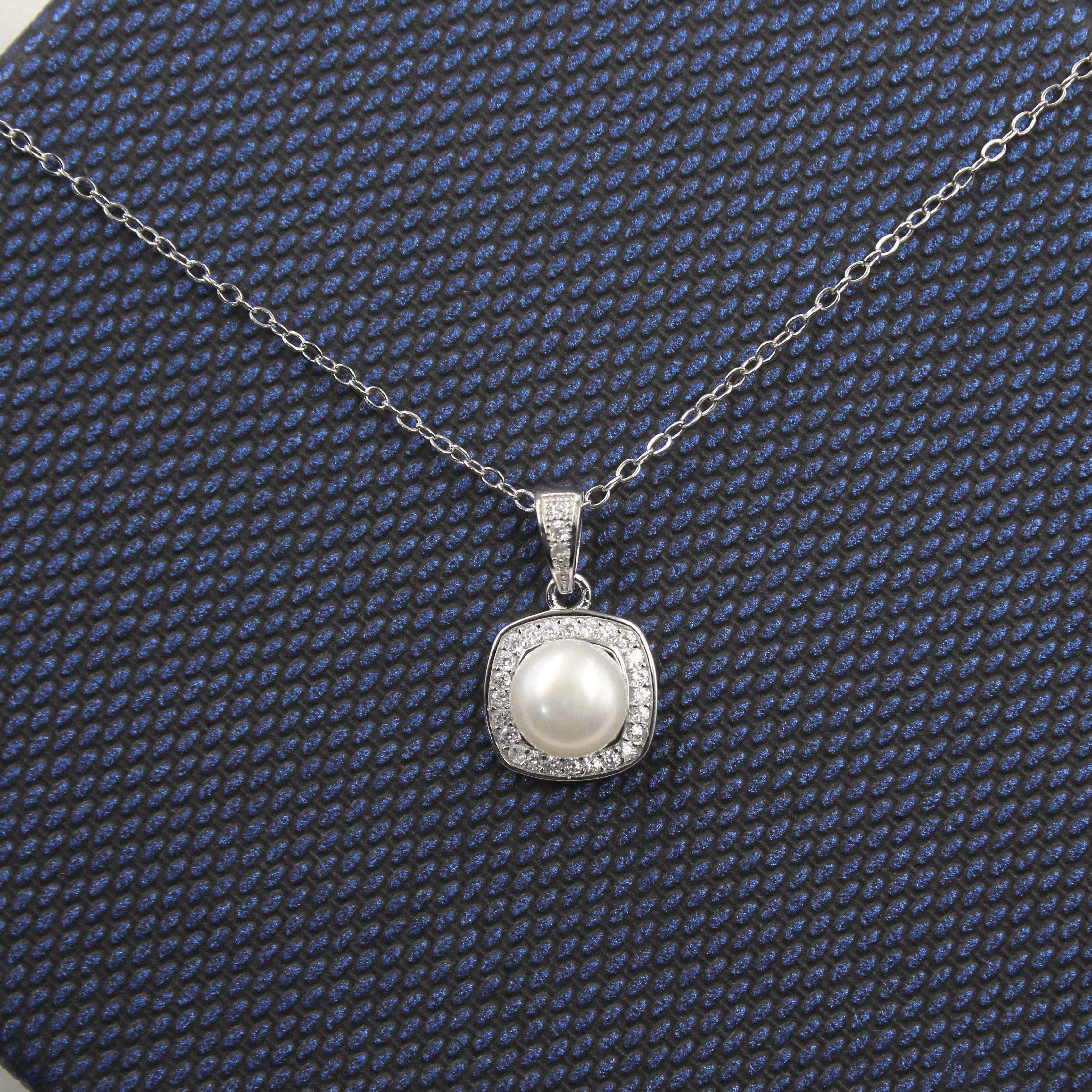 collar de plata con un colgante que presenta una perla central de color blanco, rodeada por un halo de circonias cúbicas. El colgante cuelga de una cadena de eslabones delicados y se presenta sobre una caja de regalo con textura azul oscuro, resaltando su elegancia y diseño clásico.