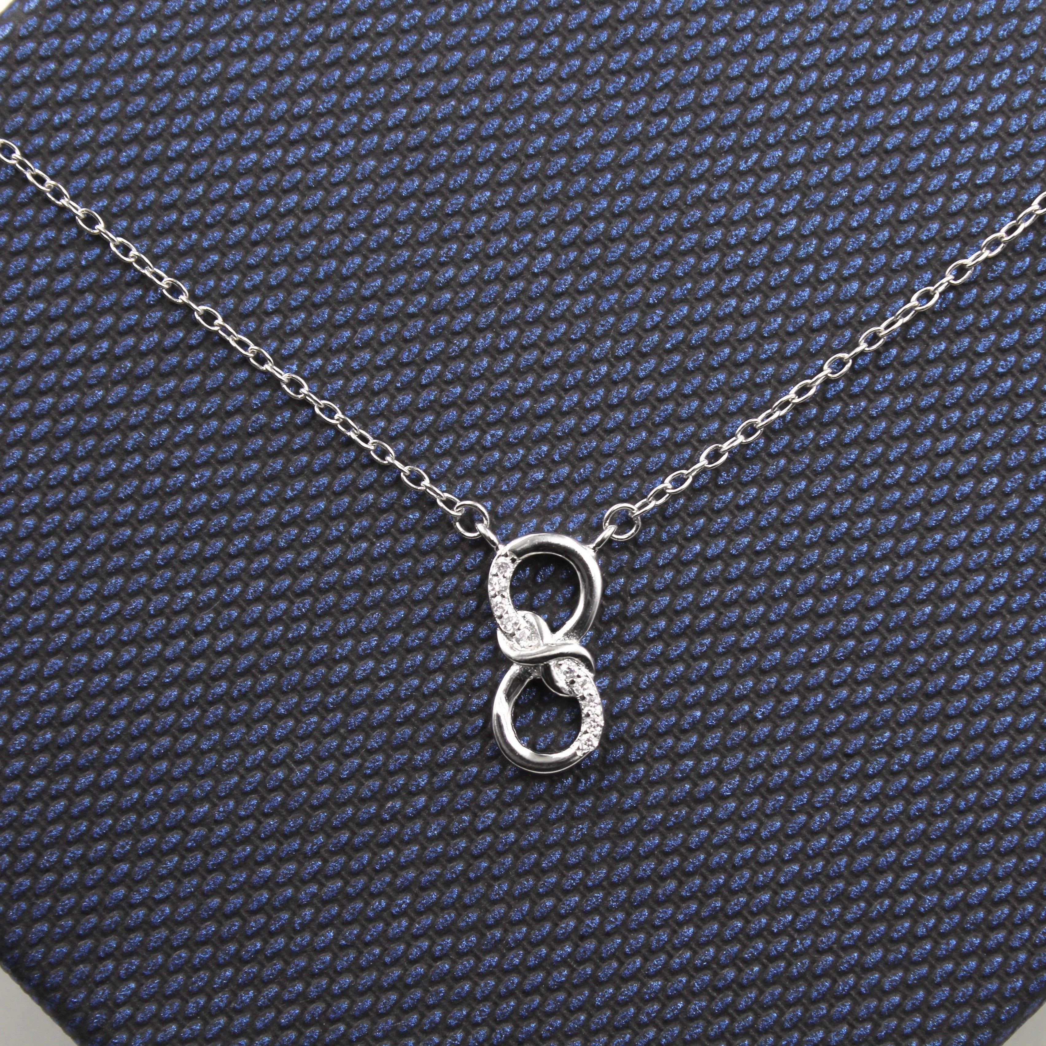 collar de plata con un colgante en forma de dos aros entrelazados sobre una caja de regalo con textura azul oscuro. El aro superior está parcialmente decorado con pequeñas circonias cúbicas que agregan un toque brillante, mientras que el aro inferior es de plata lisa.