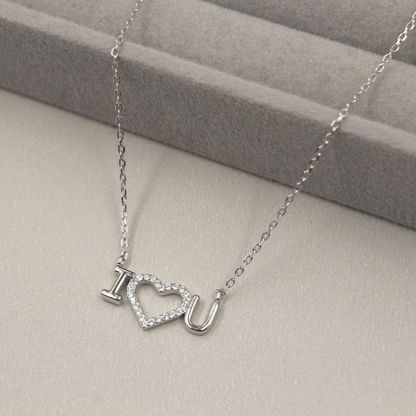 collar de plata con un colgante que forma la frase 'I ♥ U', con el corazón cubierto de pequeñas circonias cúbicas brillantes. El collar se presenta sobre un fondo gris suave con líneas horizontales, complementando el brillo del colgante.