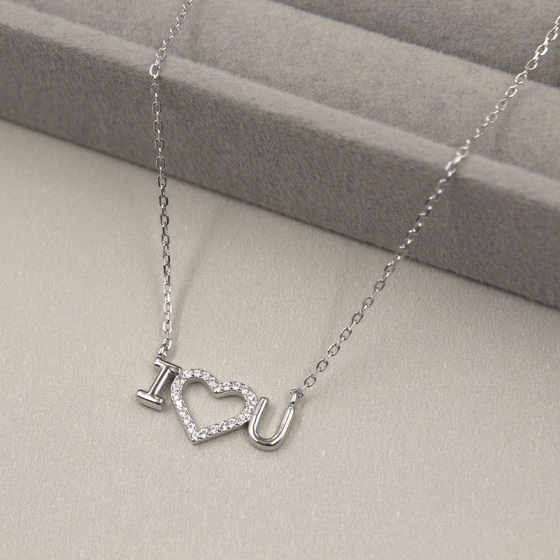 collar de plata con un colgante que forma la frase 'I ♥ U', con el corazón cubierto de pequeñas circonias cúbicas brillantes. El collar se presenta sobre un fondo gris suave con líneas horizontales, complementando el brillo del colgante.