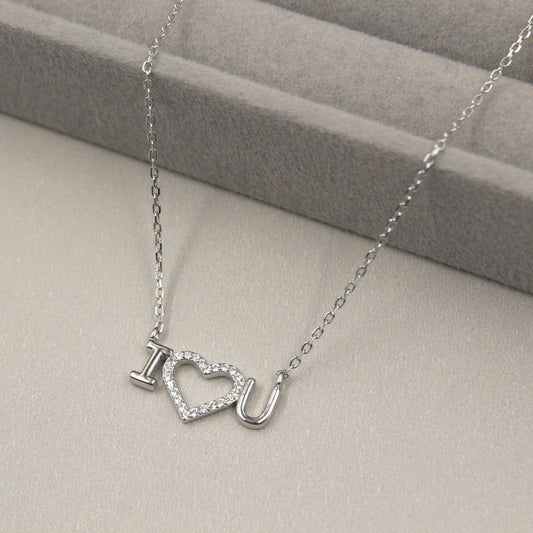 collar de plata con un colgante que forma la frase 'I ♥ U', con el corazón cubierto de pequeñas circonias cúbicas brillantes. El collar se presenta sobre un fondo gris suave con líneas horizontales, complementando el brillo del colgante.