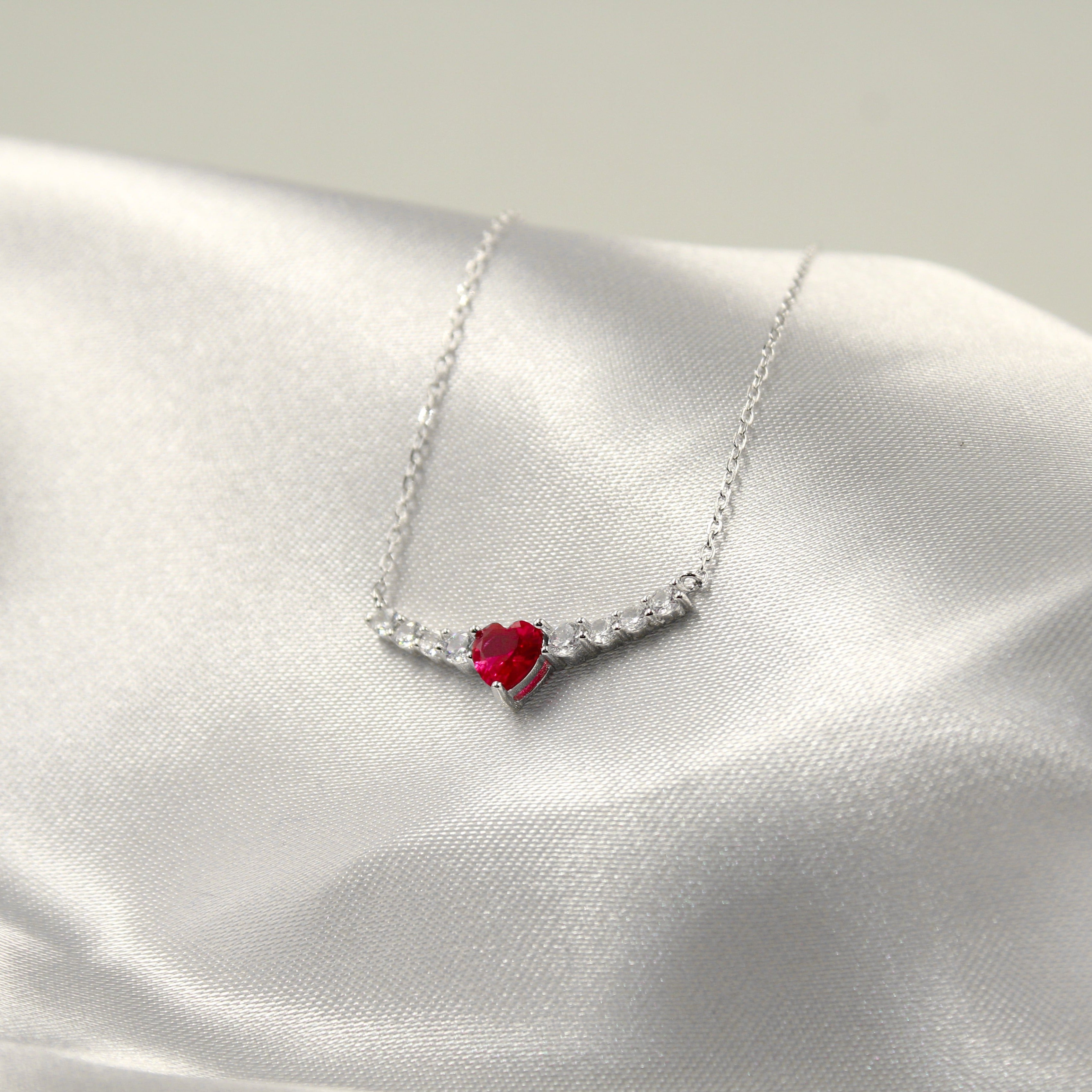 delicado collar de plata con un colgante en forma de corazón que presenta una vibrante piedra roja en el centro, rodeada por pequeñas circonias cúbicas que añaden un toque de brillo. El collar se dispone sobre una tela de satén blanco, lo que resalta su colorido y elegancia.
