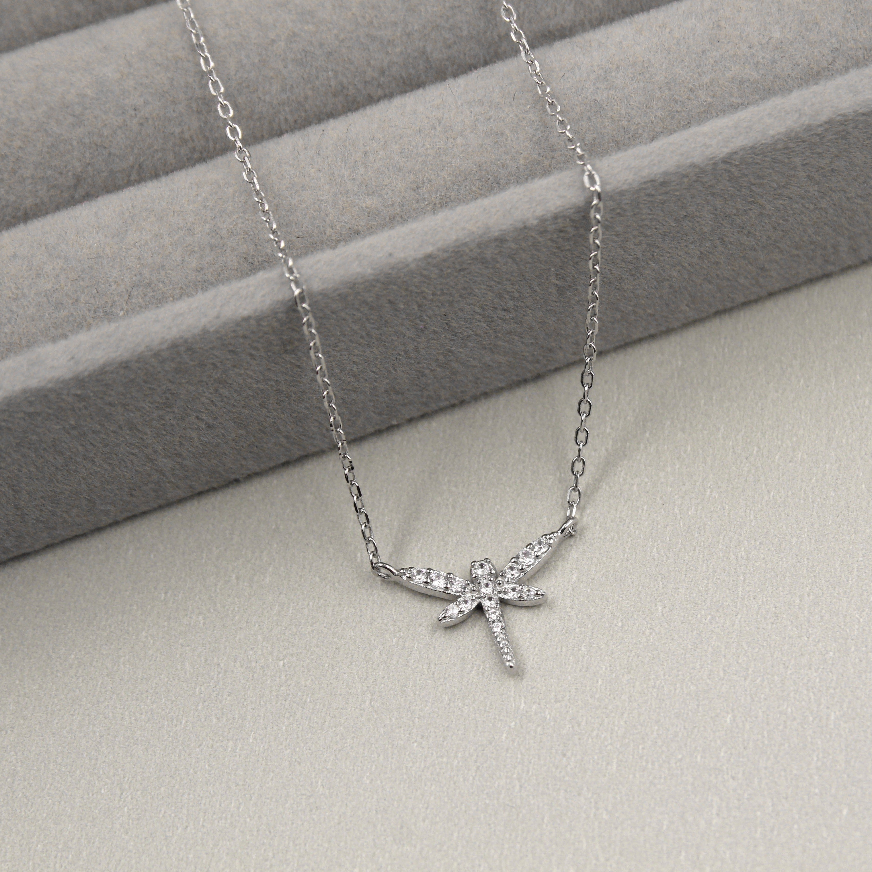 delicado collar de plata con un colgante en forma de libélula, cuyas alas y cuerpo están elegantemente adornados con pequeñas circonias cúbicas que proporcionan un brillo sutil. El colgante se presenta sobre un fondo de tela gris con líneas horizontales suaves.
