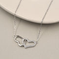 collar de plata con dos colgantes de corazón entrelazados. Uno de los corazones es liso y de plata sólida, mientras que el otro está cubierto con circonias cúbicas brillantes, ofreciendo un contraste elegante. El collar descansa sobre una placa de cerámica de color claro que realza su diseño y brillo.