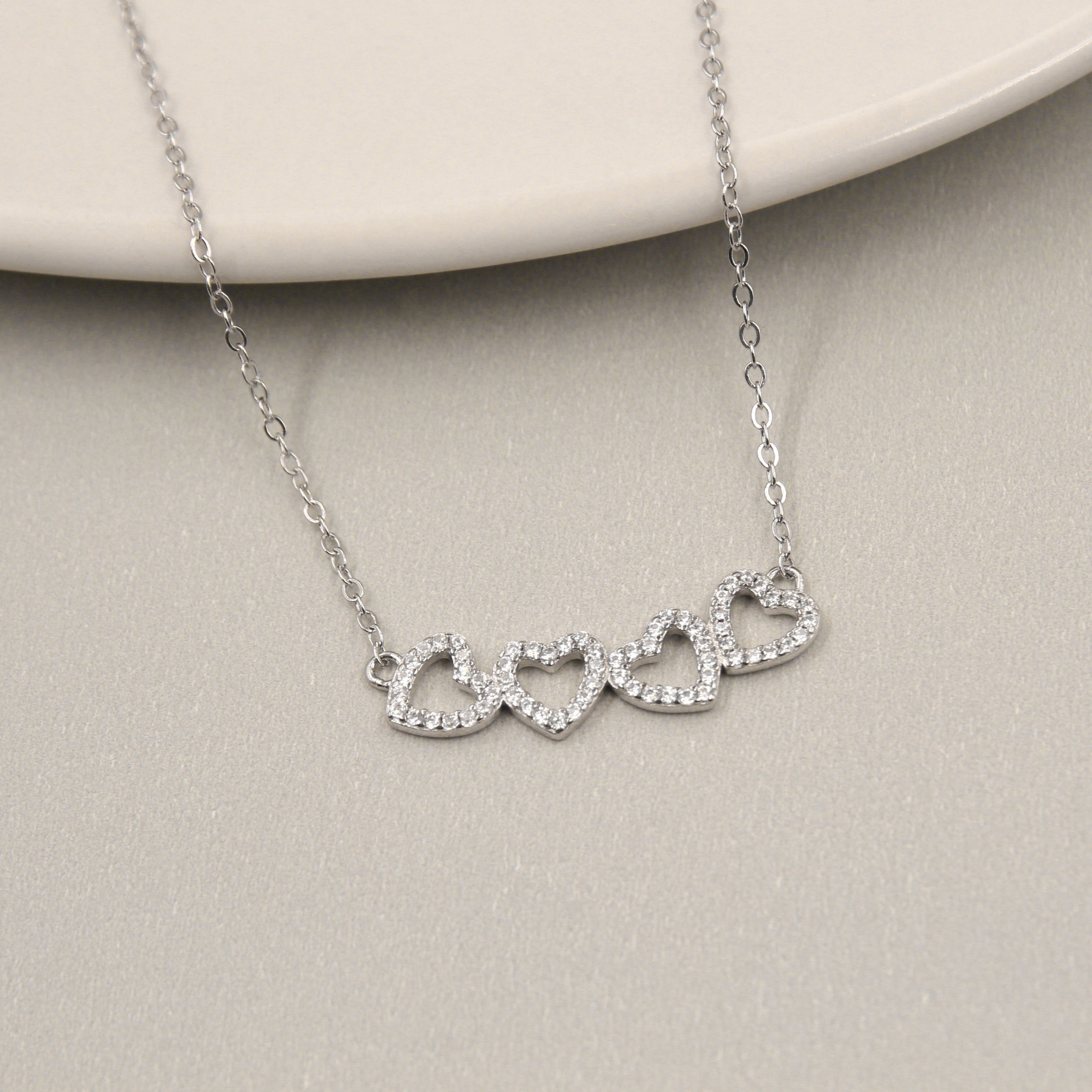  delicado collar de plata con un diseño de cuatro corazones entrelazados. Cada corazón está contorneado por pequeñas circonias cúbicas que brillan intensamente. El collar está dispuesto sobre un plato de cerámica de color claro, añadiendo un contraste suave y elegante al brillante metal.