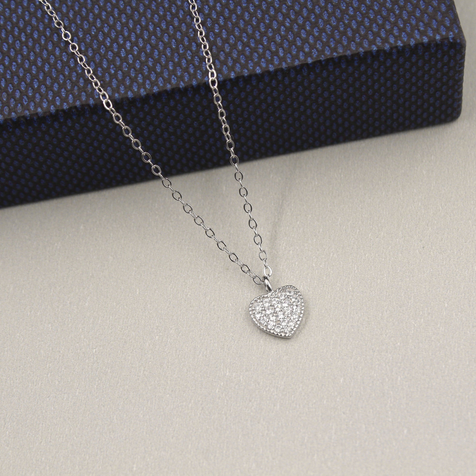 elegante collar de plata con un colgante en forma de corazón completamente cubierto de circonias cúbicas brillantes. El colgante está suspendido de una cadena de eslabones finos, presentado sobre una caja de regalo de color azul oscuro con textura, destacando su brillo y diseño romántico.