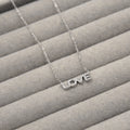 collar de plata con la palabra 'LOVE' como colgante, cada letra decorada con pequeñas circonias cúbicas que añaden un brillo sutil. El colgante está cuidadosamente colocado sobre un fondo de terciopelo gris que resalta su diseño elegante y mensaje de amor.