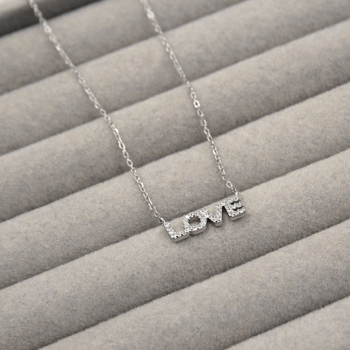 collar de plata con la palabra 'LOVE' como colgante, cada letra decorada con pequeñas circonias cúbicas que añaden un brillo sutil. El colgante está cuidadosamente colocado sobre un fondo de terciopelo gris que resalta su diseño elegante y mensaje de amor.