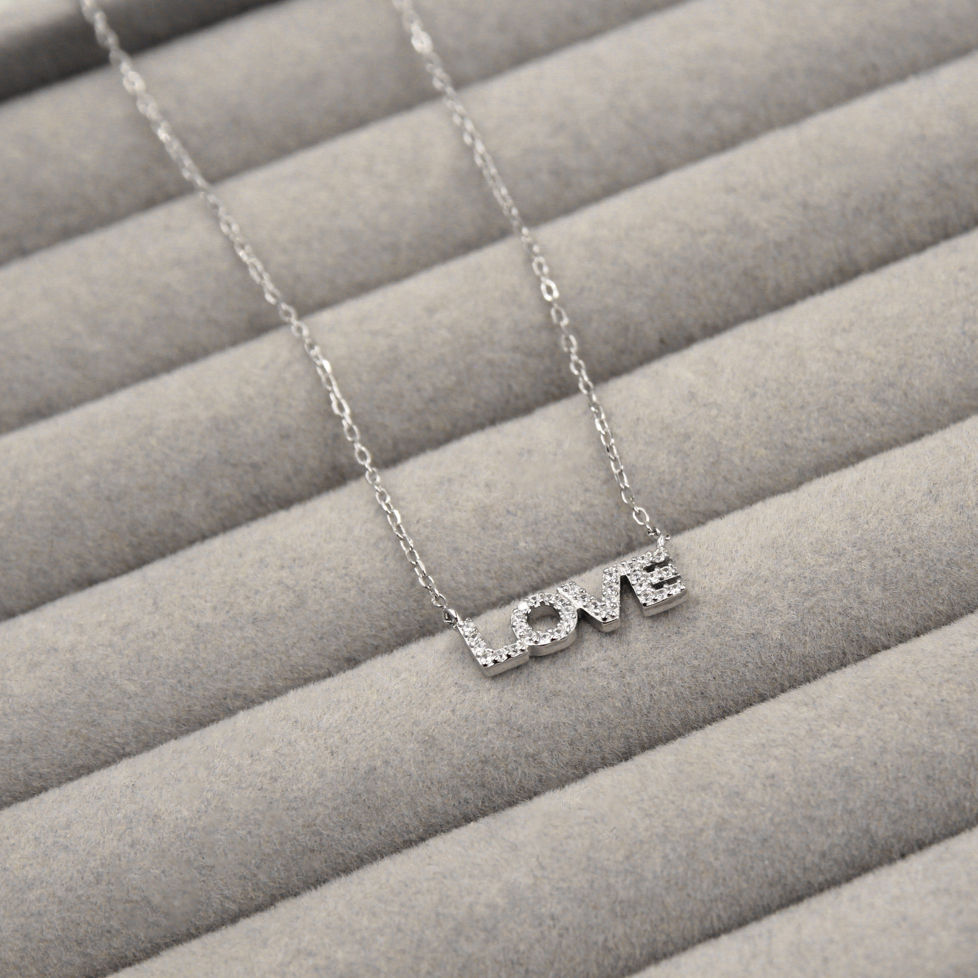 collar de plata con la palabra 'LOVE' como colgante, cada letra decorada con pequeñas circonias cúbicas que añaden un brillo sutil. El colgante está cuidadosamente colocado sobre un fondo de terciopelo gris que resalta su diseño elegante y mensaje de amor.