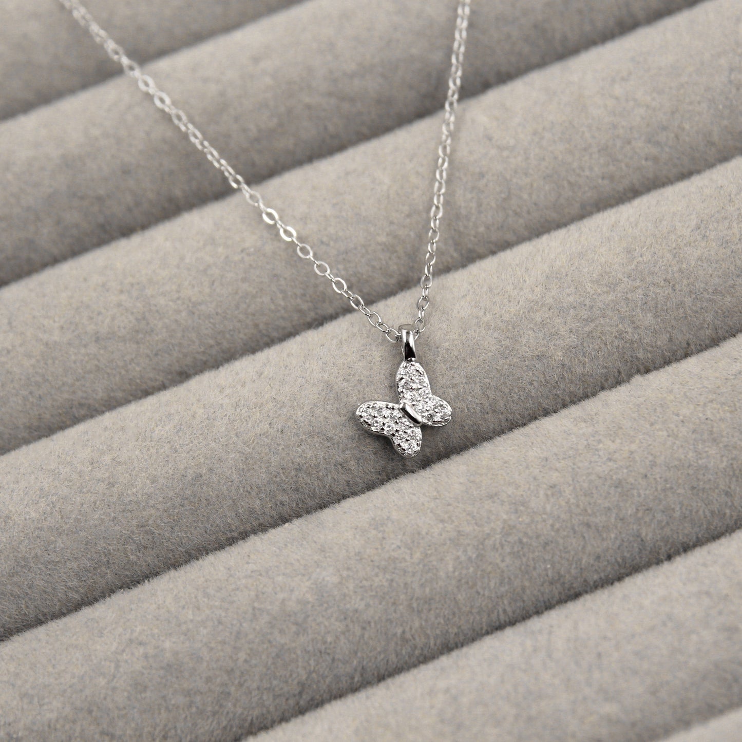 elegante collar de plata con un colgante de mariposa, cuyas alas están embellecidas con circonias cúbicas brillantes. El collar se encuentra sobre un fondo de tela gris con líneas horizontales suaves, lo que resalta la delicadeza del diseño.