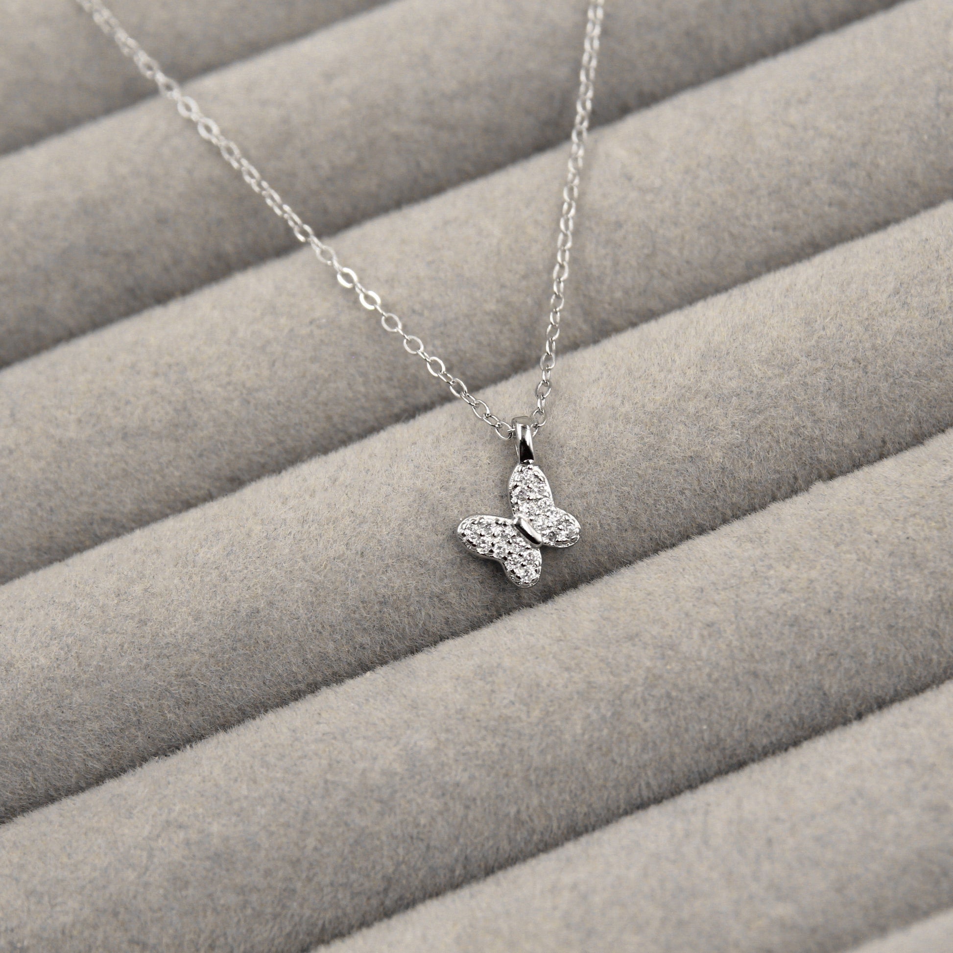 elegante collar de plata con un colgante de mariposa, cuyas alas están embellecidas con circonias cúbicas brillantes. El collar se encuentra sobre un fondo de tela gris con líneas horizontales suaves, lo que resalta la delicadeza del diseño.
