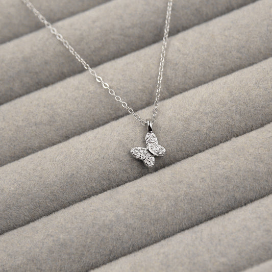 elegante collar de plata con un colgante de mariposa, cuyas alas están embellecidas con circonias cúbicas brillantes. El collar se encuentra sobre un fondo de tela gris con líneas horizontales suaves, lo que resalta la delicadeza del diseño.