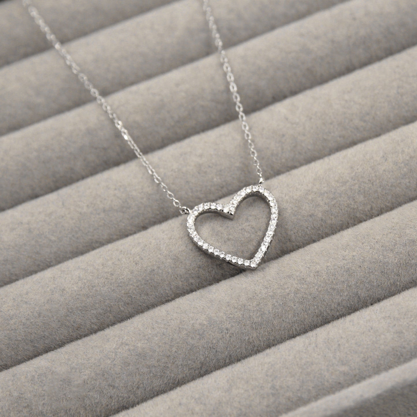 elegante collar de plata con un colgante en forma de corazón abierto, delicadamente adornado con pequeñas circonias cúbicas que cubren todo el contorno. El colgante cuelga de una cadena de eslabones finos y se presenta sobre un fondo de tela gris con líneas horizontales.