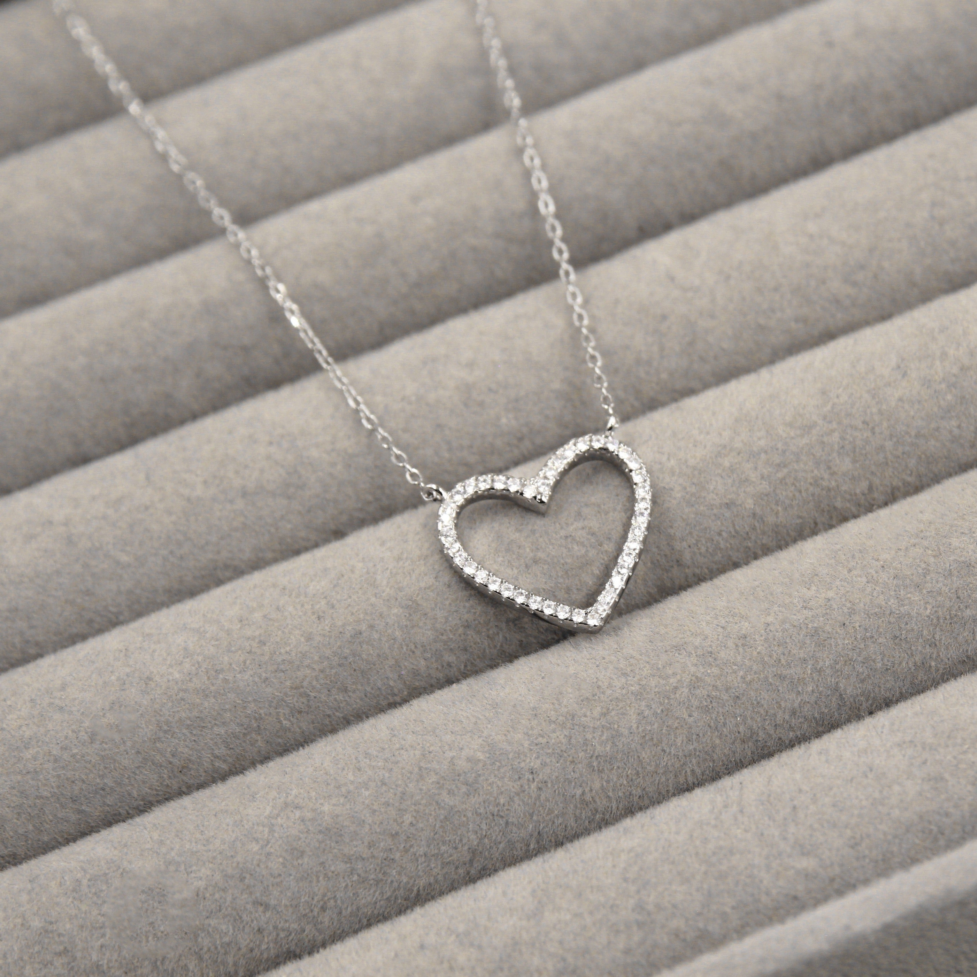 elegante collar de plata con un colgante en forma de corazón abierto, delicadamente adornado con pequeñas circonias cúbicas que cubren todo el contorno. El colgante cuelga de una cadena de eslabones finos y se presenta sobre un fondo de tela gris con líneas horizontales.