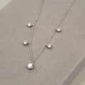 collar de plata con un diseño en cascada de cinco piedras circulares de circonia cúbica, distribuidas a lo largo de la cadena con espaciado uniforme. Cada piedra está montada en una simple pero elegante montura que captura la luz, brillando intensamente sobre un fondo de terciopelo gris.