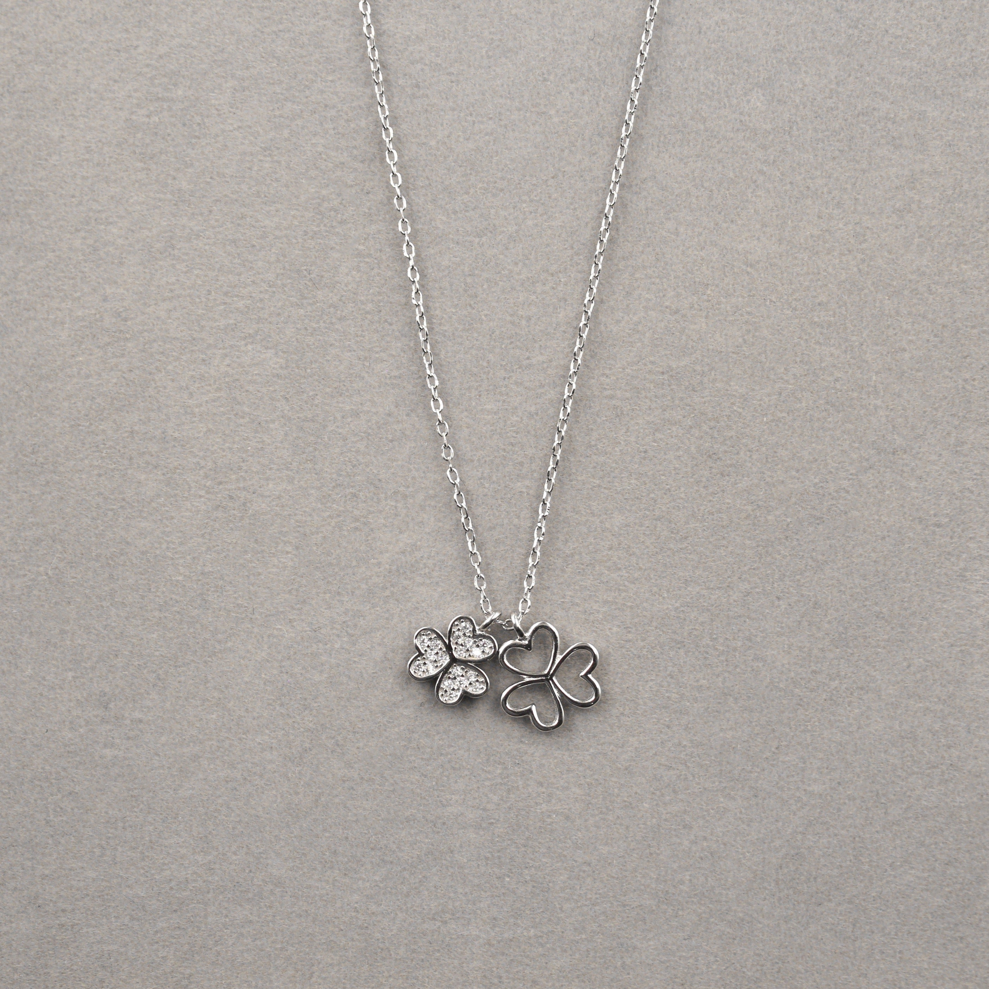  collar de plata con un colgante compuesto por dos tréboles de tres hojas, colocado sobre un fondo gris. Dos de los tréboles son de plata sólida y el segundo, situado en el centro, está decorado con pequeñas circonias cúbicas que imitan el brillo de diamantes. 