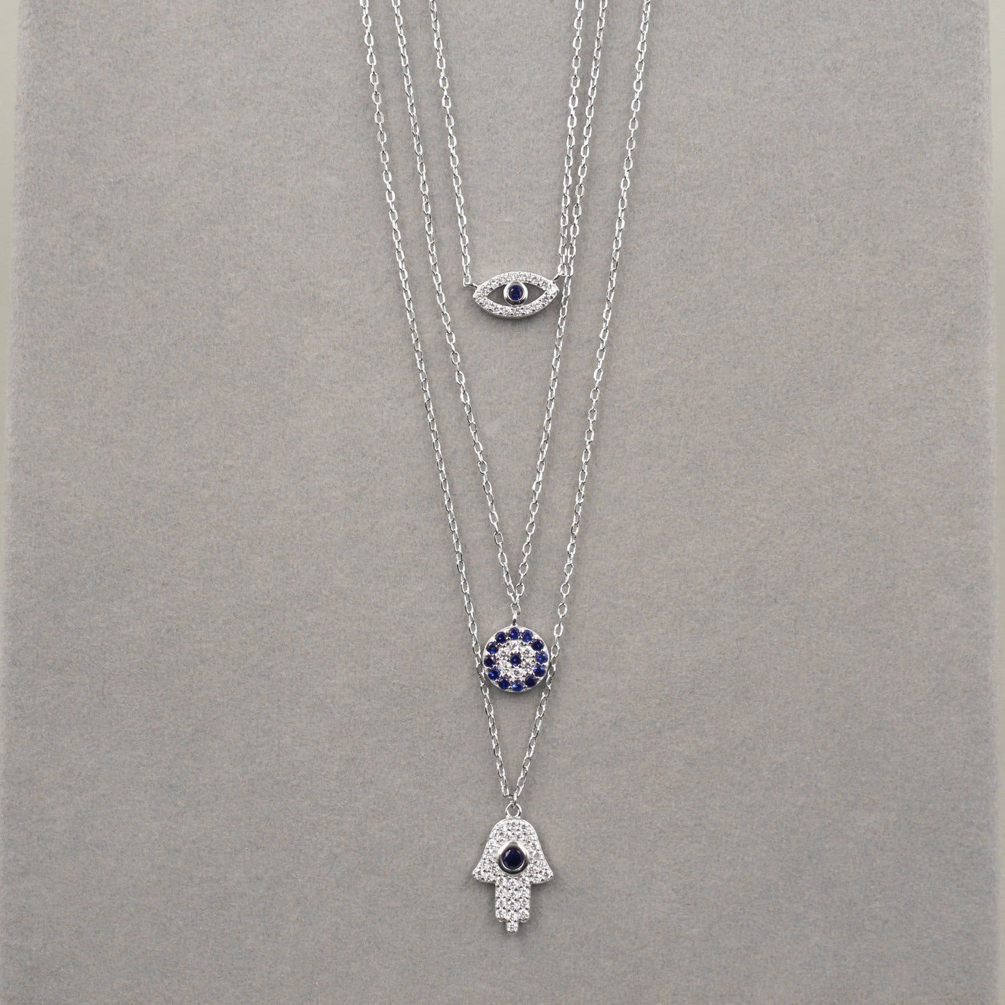 res collares de plata dispuestos en capas, cada uno con un colgante diferente. El colgante superior es un ojo con una piedra azul central, el del medio presenta un diseño circular con pequeñas piedras azules, y el inferior es una mano de Hamsa adornada con circonias cúbicas. Los collares están sobre un fondo de terciopelo gris, destacando su elegancia y diseño simbólico.
