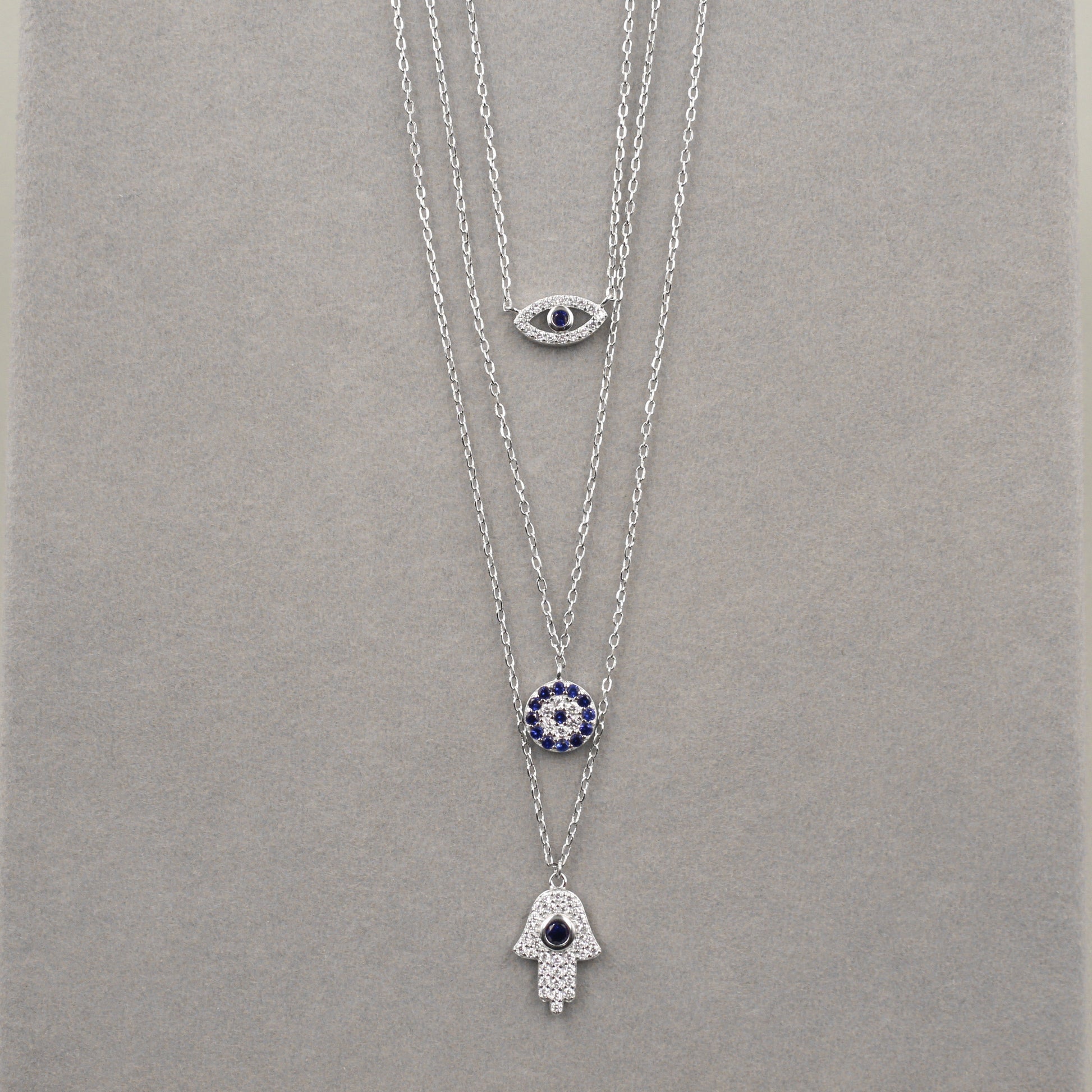 res collares de plata dispuestos en capas, cada uno con un colgante diferente. El colgante superior es un ojo con una piedra azul central, el del medio presenta un diseño circular con pequeñas piedras azules, y el inferior es una mano de Hamsa adornada con circonias cúbicas. Los collares están sobre un fondo de terciopelo gris, destacando su elegancia y diseño simbólico.