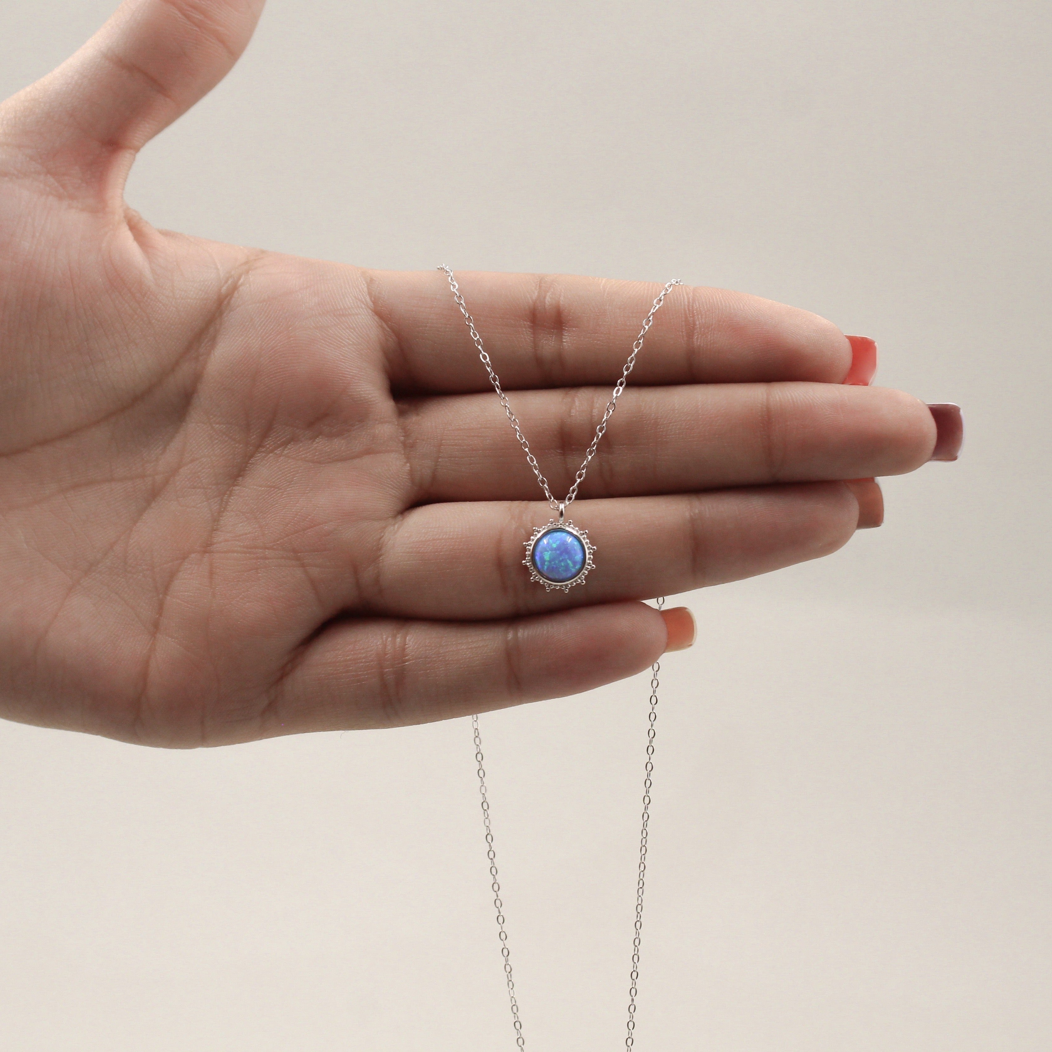 una mano sosteniendo un collar de plata con un colgante redondo que alberga una piedra de ópalo azul vibrante, rodeada por un halo de pequeñas circonias cúbicas. El diseño capta la luz, mostrando destellos de colores dentro del ópalo, mientras la cadena delicada se extiende elegantemente desde ambos lados del colgante.