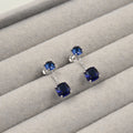 Par de aretes largos con zafiros azules cuadrados, con una piedra en la parte superior y otra en la parte inferior, unidos por una barra de plata, presentados en un estuche de joyería gris.