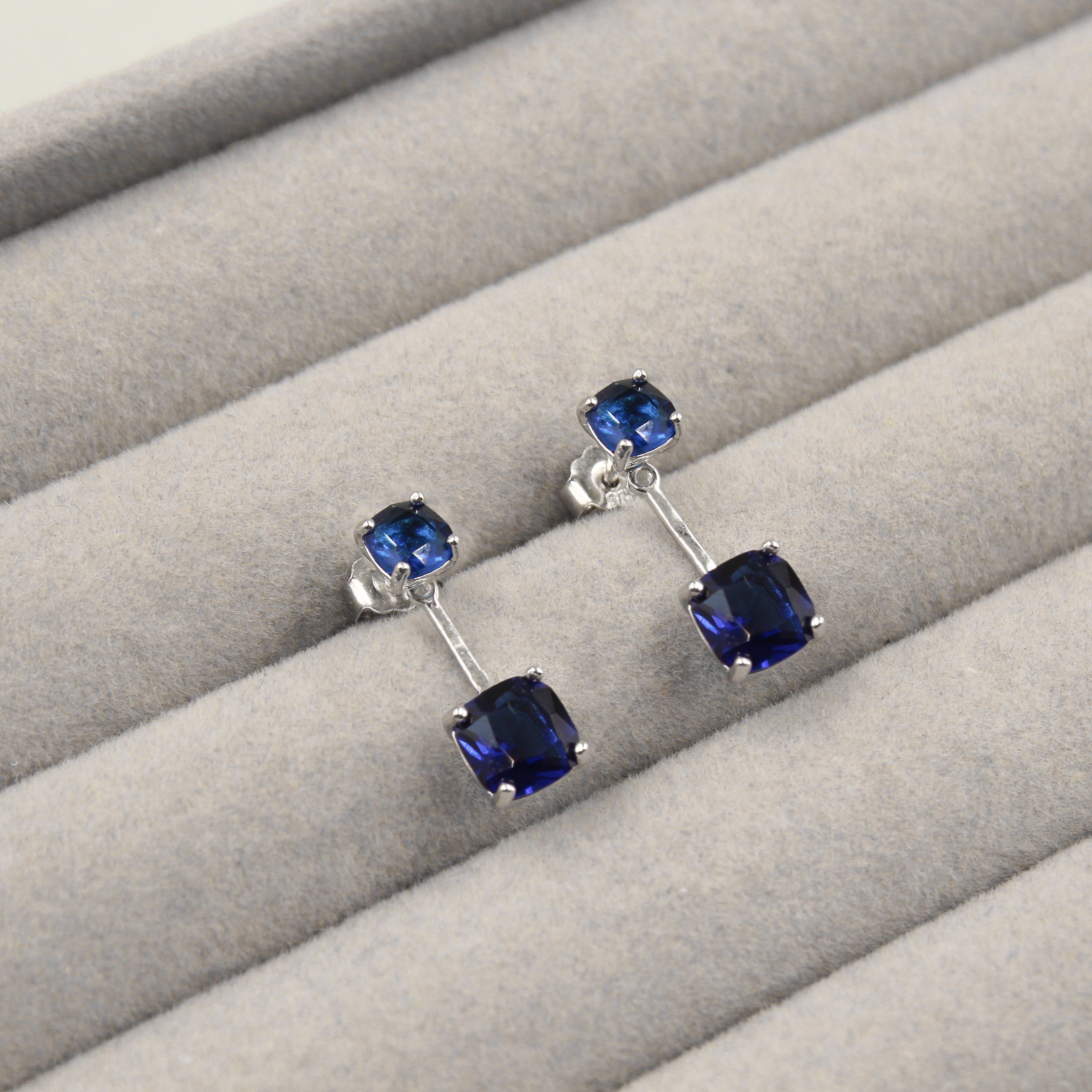 Par de aretes largos con zafiros azules cuadrados, con una piedra en la parte superior y otra en la parte inferior, unidos por una barra de plata, presentados en un estuche de joyería gris.