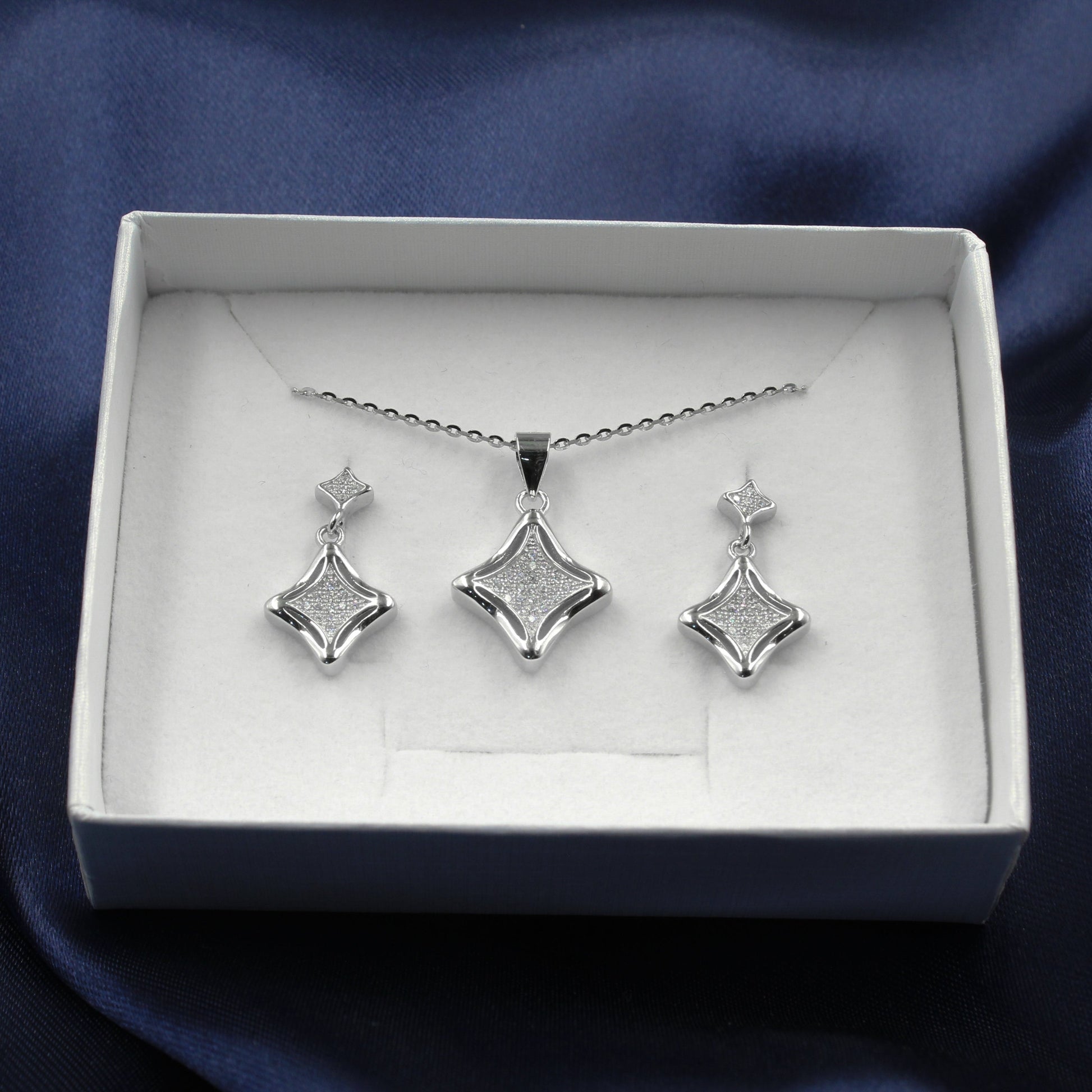 Juego de joyería en caja blanca con colgante y aretes en forma de rombo decorados con pequeños diamantes, sobre fondo de tela azul..