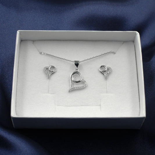 Juego de joyería en caja blanca con colgante y aretes en forma de corazón estilizado decorados con pequeños diamantes, sobre fondo de tela azul.