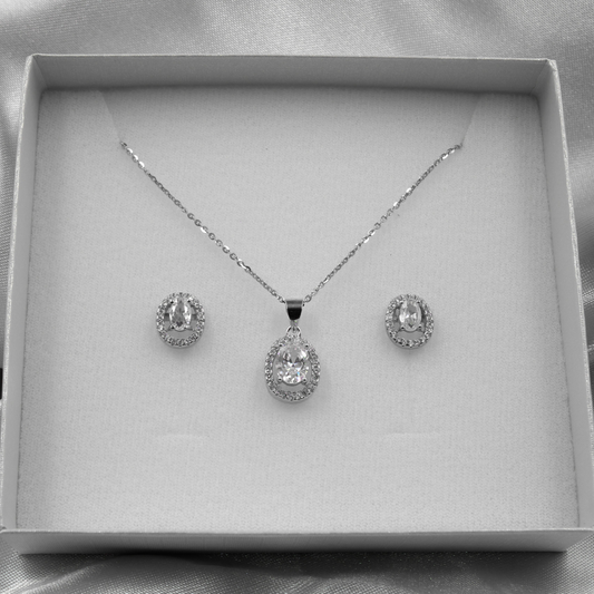 Juego de joyería en caja blanca con colgante y aretes ovalados decorados con pequeños diamantes, sobre fondo de tela plateada.