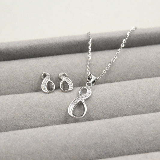 Juego de joyería con colgante y aretes en forma de infinito, decorados con pequeños diamantes, presentados en una caja de joyería gris.