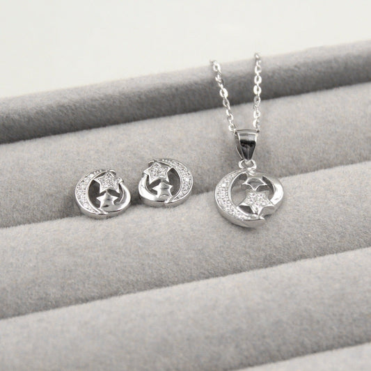 Juego de joyería con colgante y aretes en forma de luna y estrella, decorados con pequeños diamantes, presentados en una caja de joyería gris.