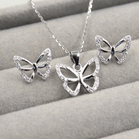 Juego de joyería con colgante y aretes en forma de mariposa, decorados con pequeños diamantes, presentados en una caja de joyería gris.